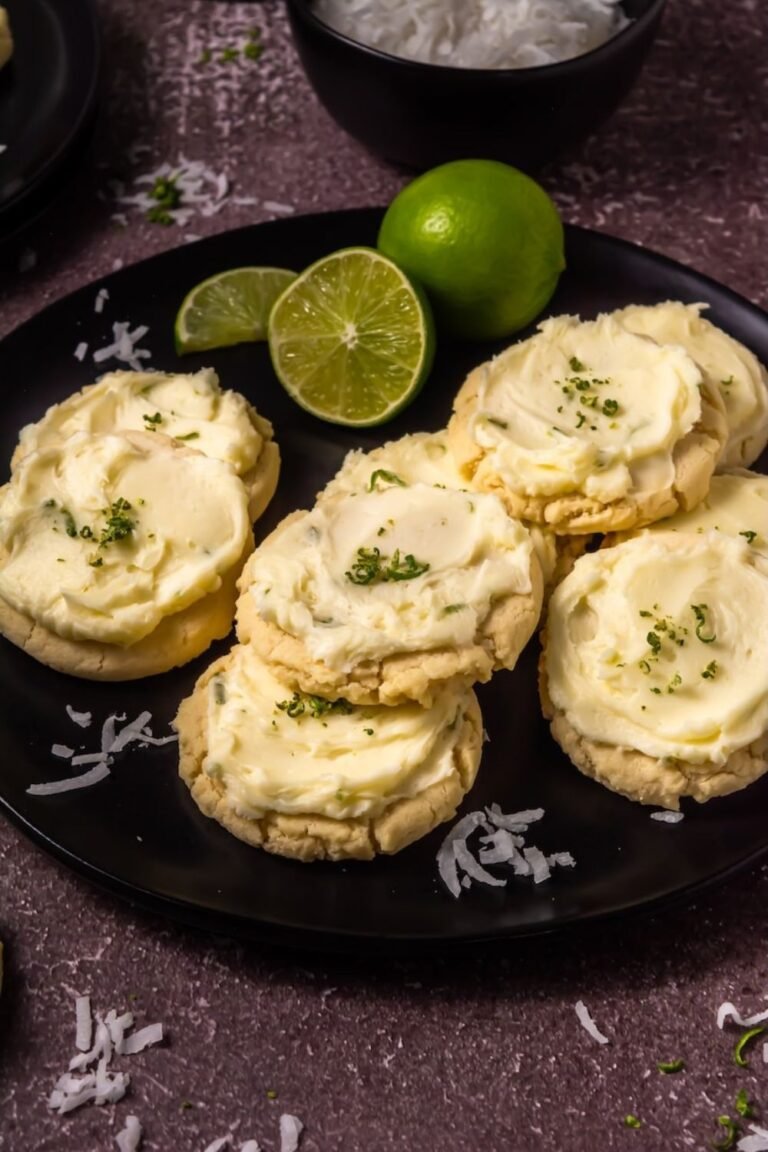 Overhead view of soft coconut lime cookies generously spread with creamy lime buttercream frosting and sprinkled with fresh lime zest, arranged on a black plate with whole and sliced limes and scattered coconut flakes in the background.