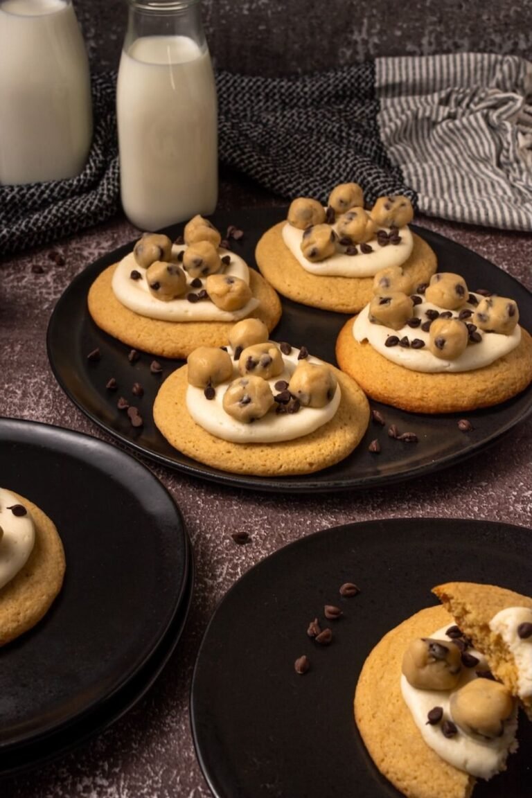 Homemade Crumbl cookie dough cookies arranged on black plates, each topped with creamy vanilla frosting, edible chocolate chip cookie dough balls, and mini chocolate chips, with bottles of milk in the background.