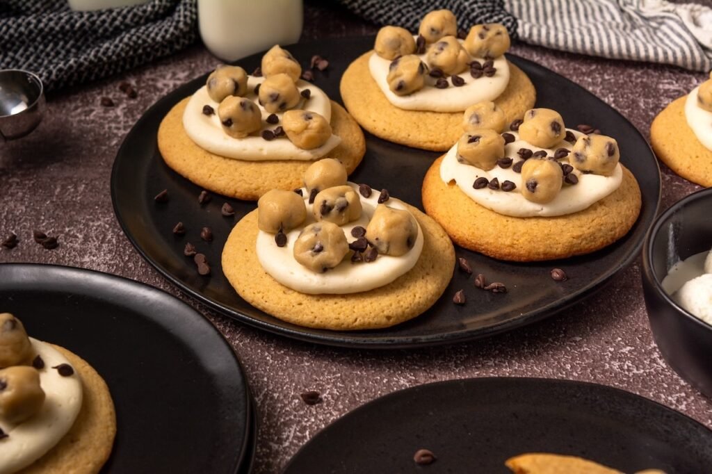 Close-up view of homemade Crumbl cookie dough cookies on a black serving plate topped with creamy vanilla frosting, edible chocolate chip cookie dough balls, and mini chocolate chips.