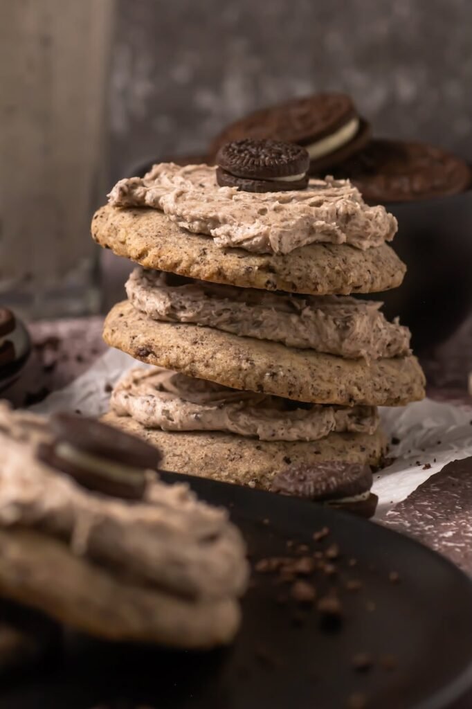 Close view of a stack of crumbl cookies and cream milkshake cookies layered with thick cookies-and-cream frosting and topped with mini Oreo cookies, showing the soft chewy cookie texture and speckled Oreo crumbs.