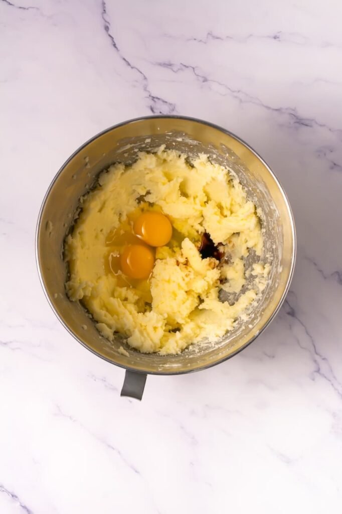 Stand mixer bowl with creamed butter and sugar as eggs and vanilla are added to begin making cookies and cream milkshake cookie dough.