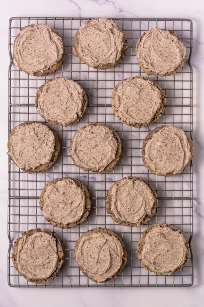 Overhead view of cookies and cream cookies spread with cookies-and-cream frosting on a wire cooling rack before adding mini Oreo toppings.