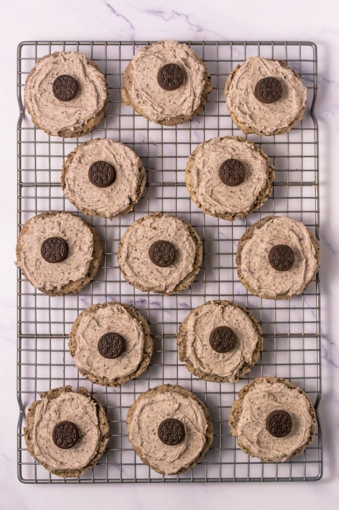 Overhead view of thick cookies and cream cookies spread with cookies-and-cream frosting and topped with mini Oreo cookies, arranged on a metal cooling rack on a marble countertop.