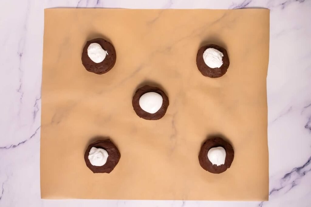 Chocolate cookie dough balls on parchment paper with frozen marshmallow fluff placed in the center before sealing the dough.