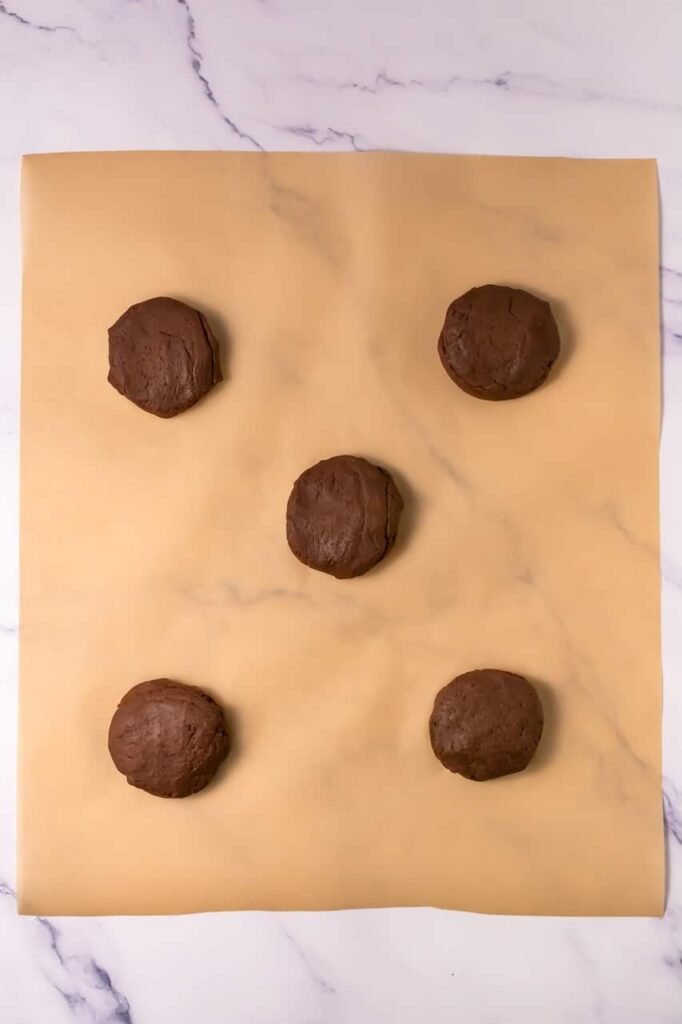 Five large chocolate cookie dough balls arranged on parchment paper, shaped and ready to bake into mallow cupcake cookies.