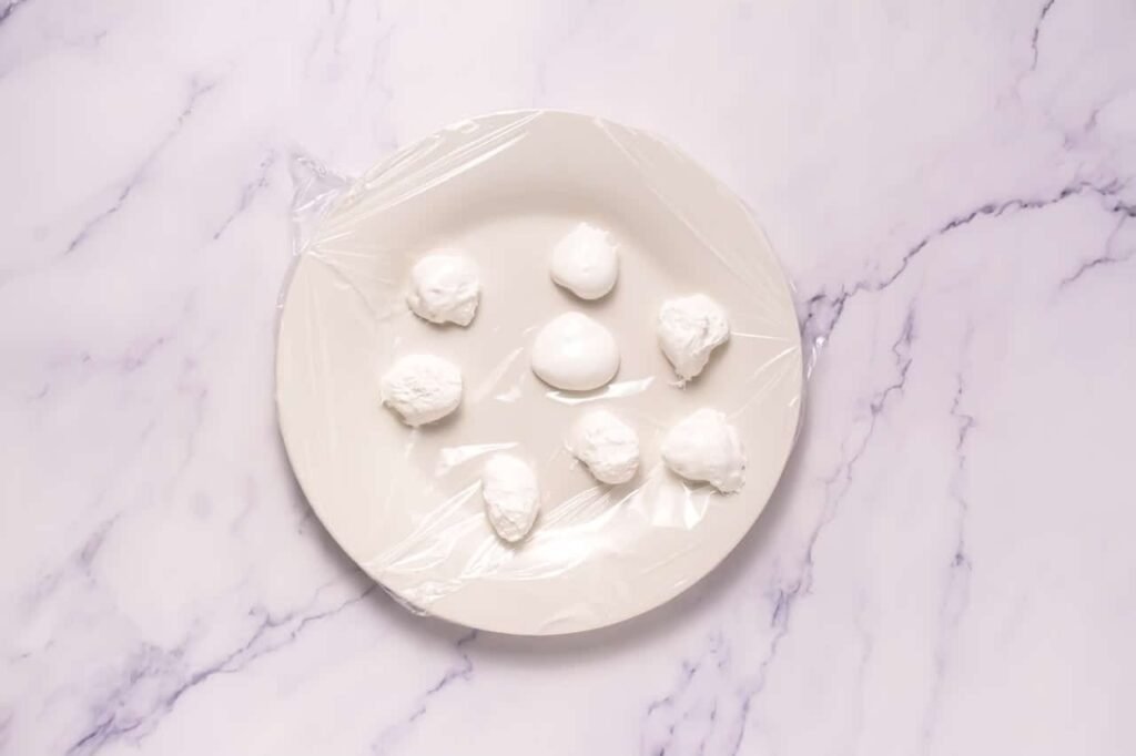 Plate lined with plastic wrap holding small scoops of marshmallow fluff that are being frozen before filling chocolate mallow cupcake cookies.