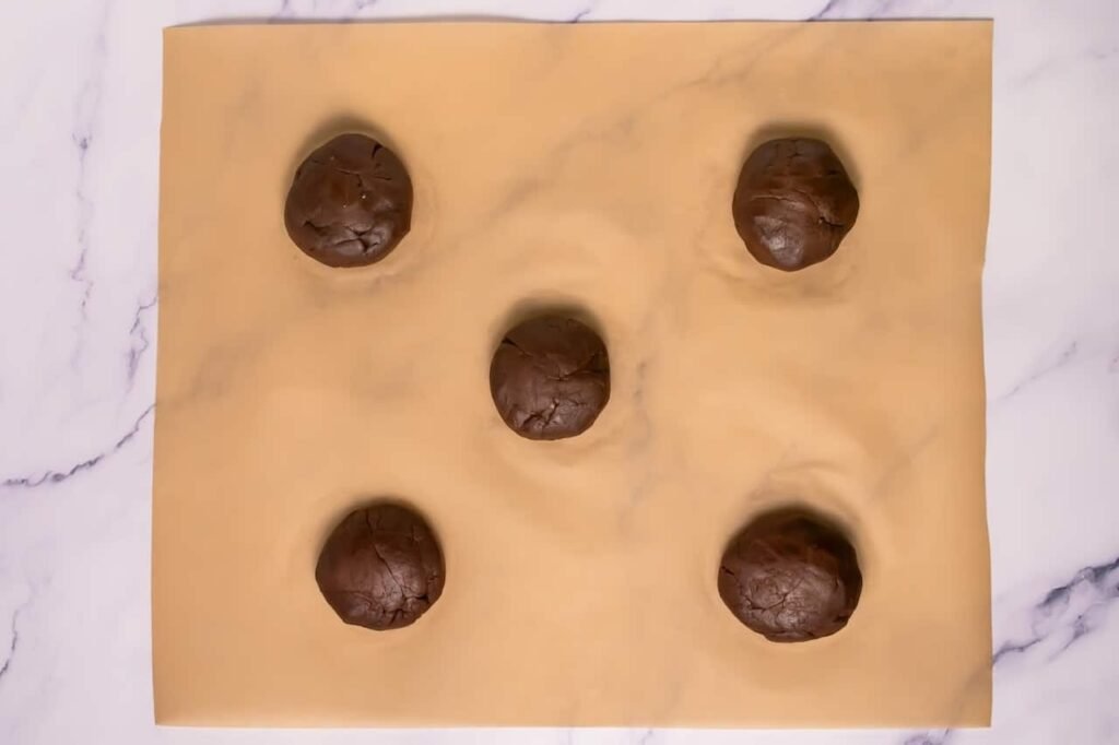 Chocolate cookie dough balls on parchment paper with marshmallow filling sealed inside, shaped and ready to bake.