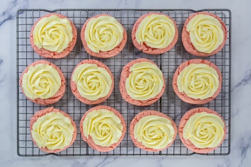 Overhead view of strawberry cookies on a cooling rack topped with piped cream cheese frosting swirls, showing bakery-style presentation before adding crumble topping.