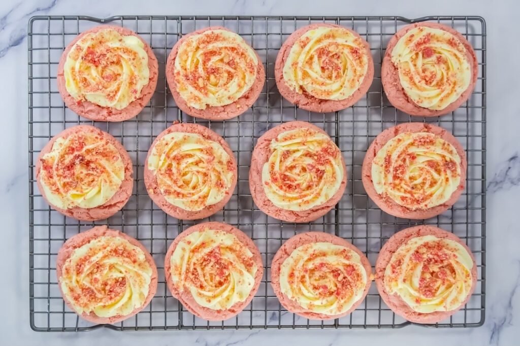 Overhead view of strawberry shortcake cookies on a cooling rack topped with piped frosting and sprinkled strawberry crumble, showing finished bakery-style cookies.