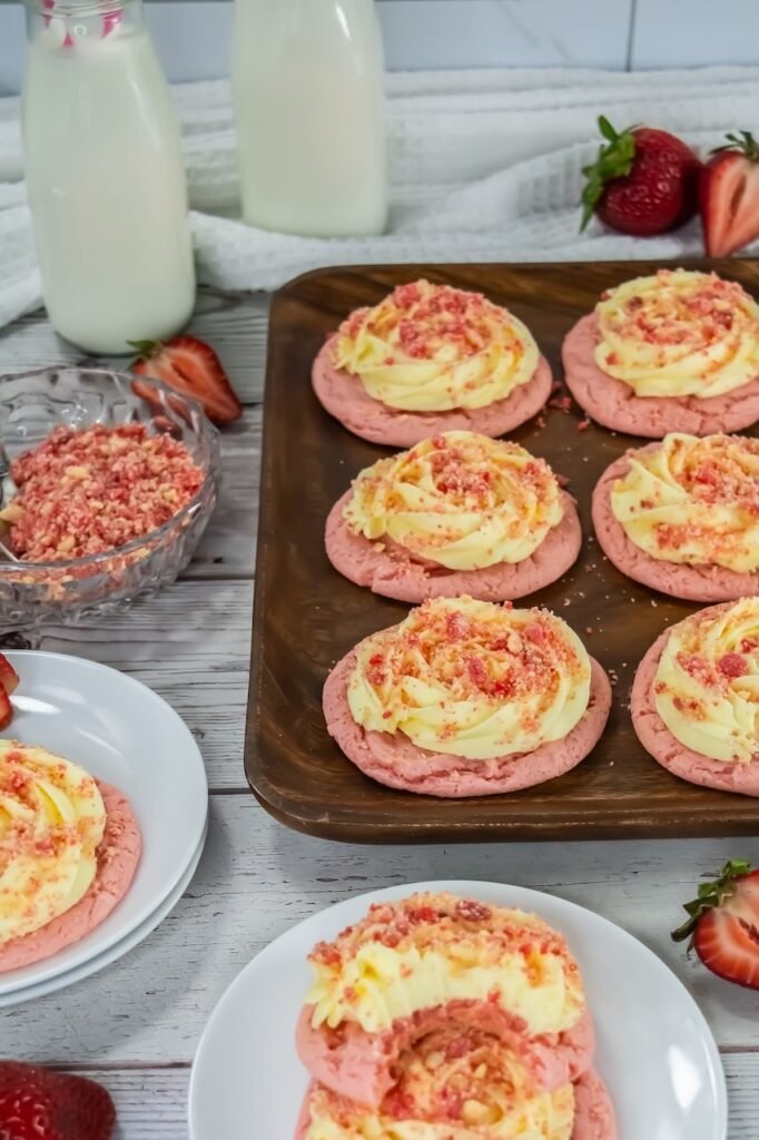 Strawberry shortcake cookies served on a wooden tray with frosting and crumble topping, alongside milk bottles and fresh strawberries on a table.