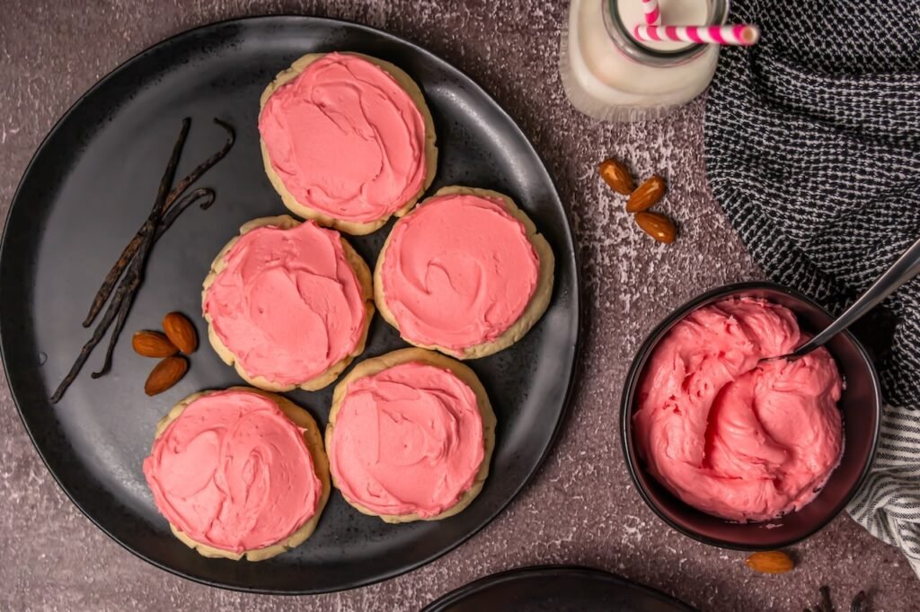 Close-up of soft sugar cookies with pink frosting arranged on black plates, styled with milk and almonds in the background for serving.