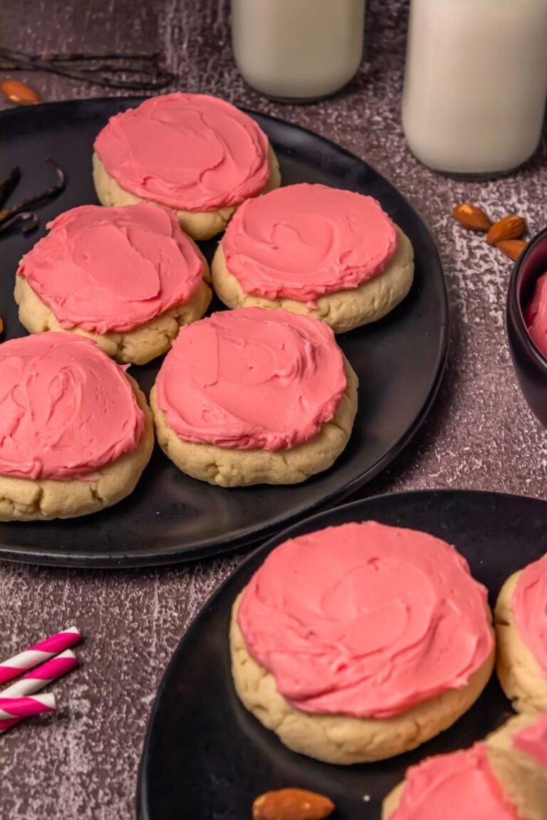 Close-up of soft sugar cookies with pink frosting arranged on black plates, styled with milk and almonds in the background for serving.