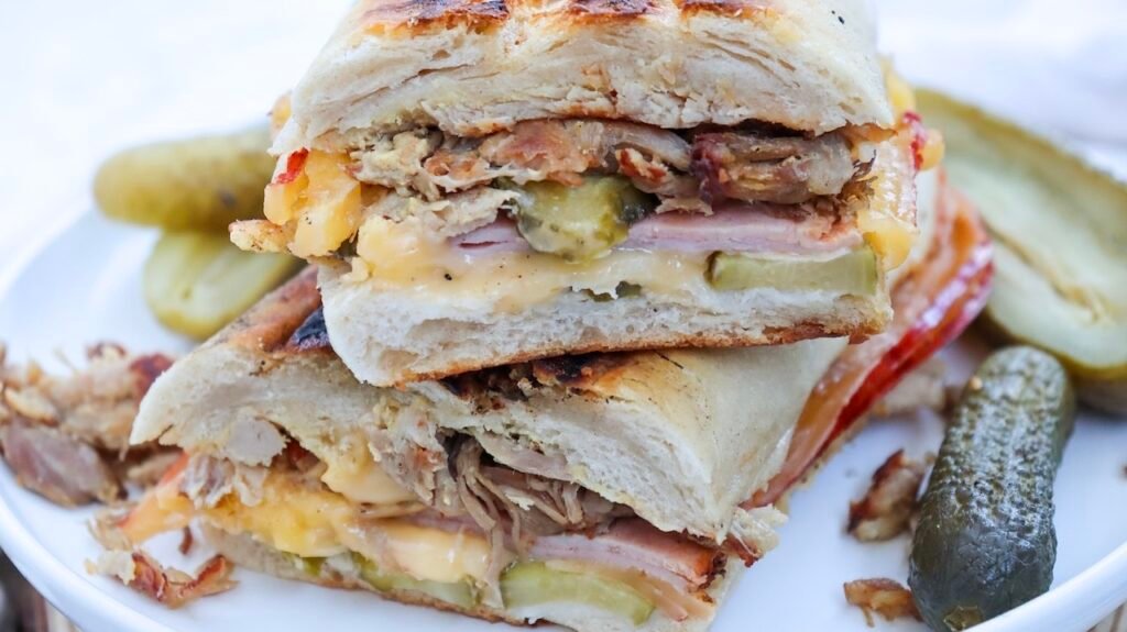 Close-up cross-section of a pressed Cuban sandwich showing layers of roast pork, ham, melted Swiss cheese, and sliced pickles inside golden toasted bread on a white plate.