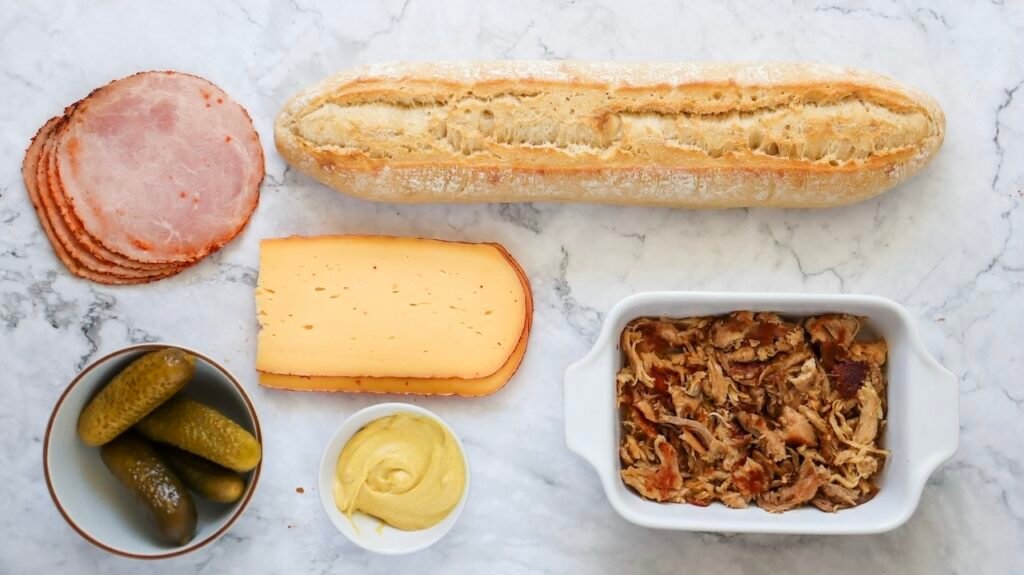 Overhead view of Cuban sandwich ingredients arranged on a marble surface, including sliced roast pork, ham, Swiss cheese, dill pickles, yellow mustard, and a loaf of Cuban bread ready for assembly.