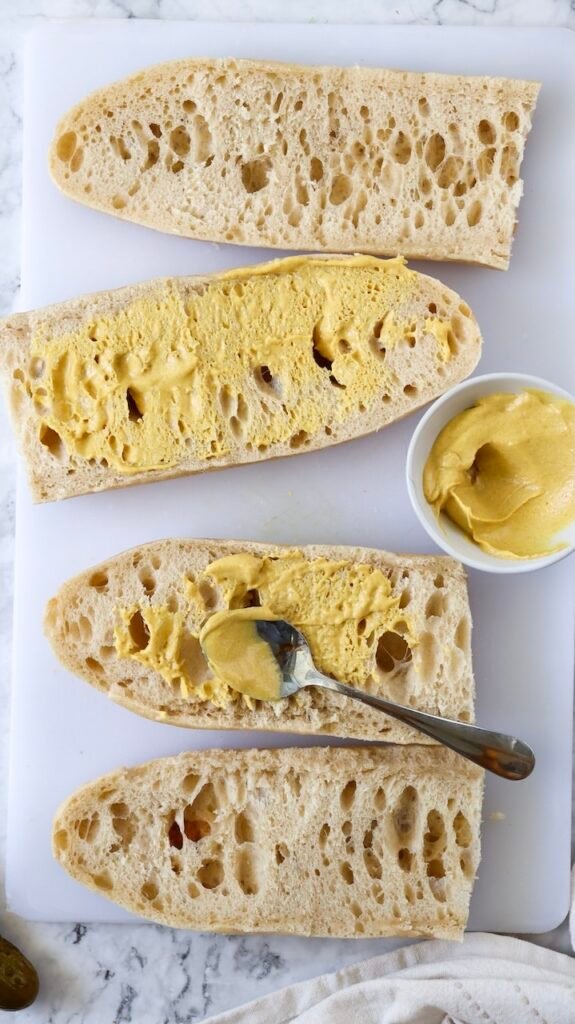 Overhead view of sliced Cuban bread on a cutting board with yellow mustard spread evenly on the inside, a spoon resting on one half, and a small bowl of mustard nearby, showing the preparation step before assembling a Cuban sandwich.