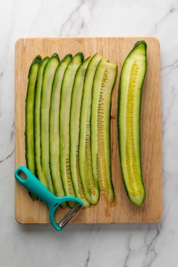 Thin cucumber ribbons sliced lengthwise on a wooden cutting board with a vegetable peeler, prepared for smoked salmon cucumber rolls.