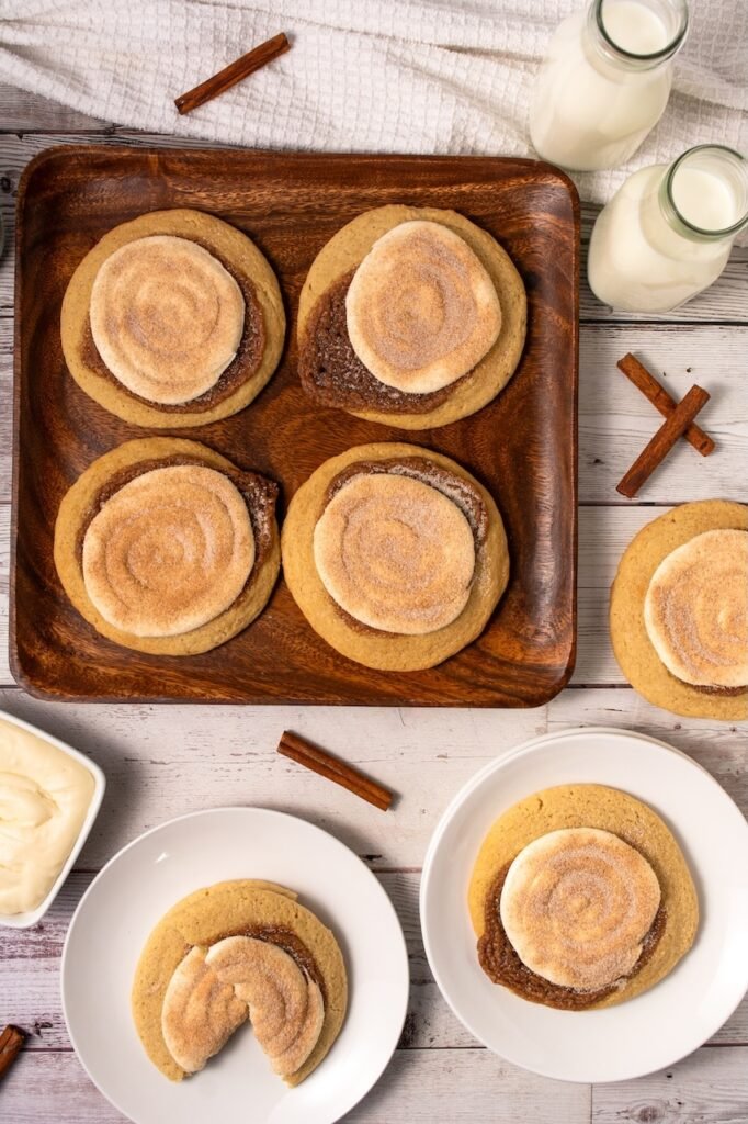 Overhead view of several Crumbl cinnamon swirl cookies arranged on a wooden tray and white plates, with one cookie broken open to show the soft interior, surrounded by cinnamon sticks, frosting, and small bottles of milk.