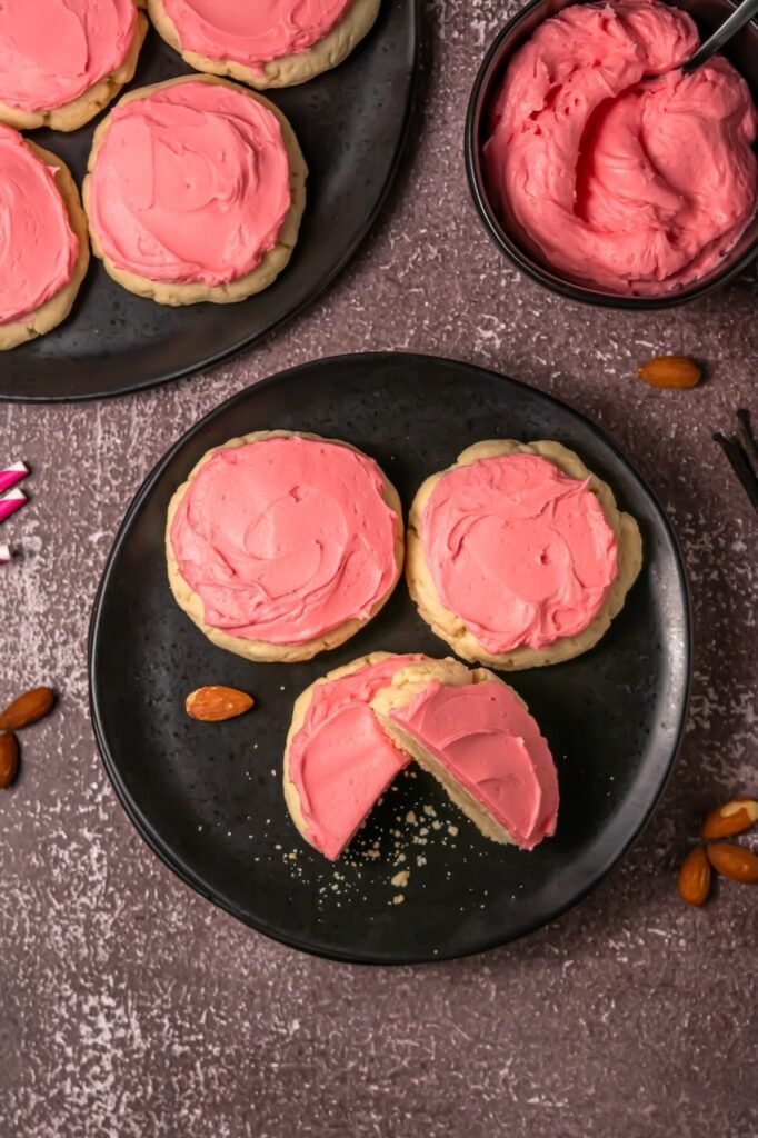 Overhead view of frosted sugar cookies on black plates, including one cookie cut in half to show the soft interior and pink frosting.