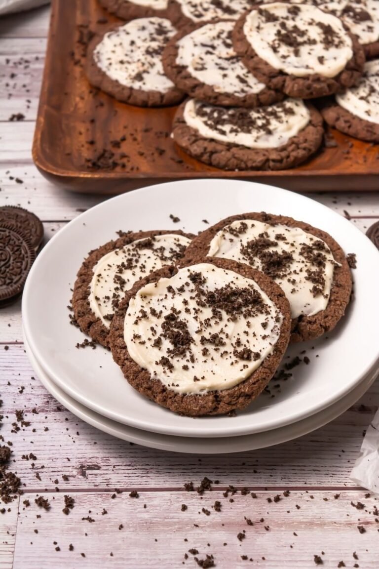 Close-up of crumbl chocolate Oreo cookies with smooth frosting and Oreo crumbs served on a white plate, with more cookies in the background on a tray.