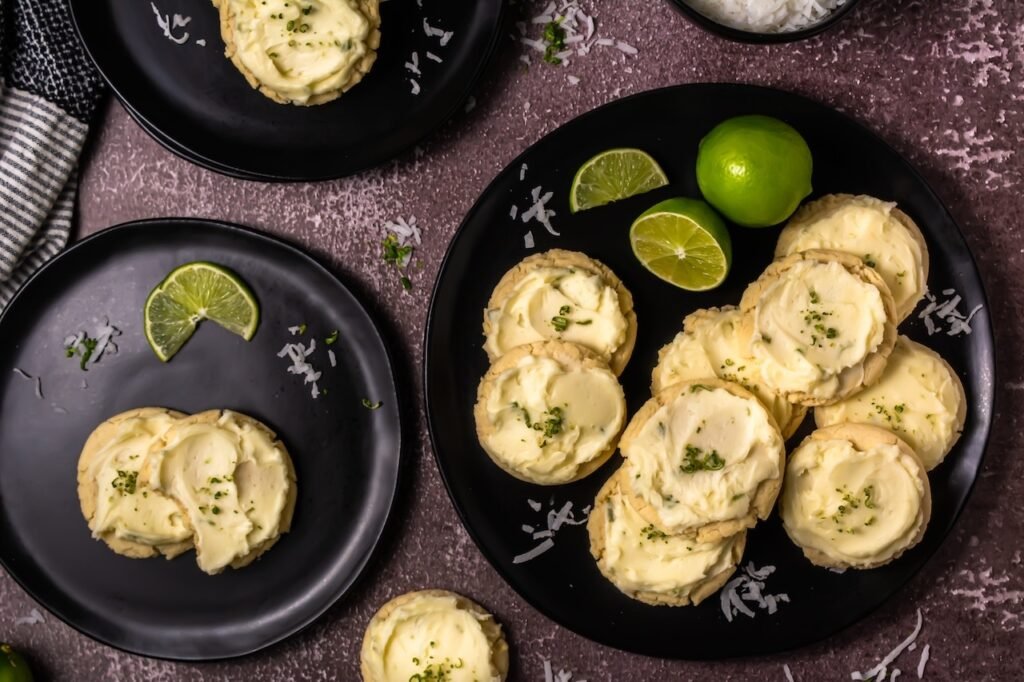 Overhead view of thick coconut lime cookies topped with creamy lime frosting and lime zest served on black plates with fresh lime slices and coconut flakes.