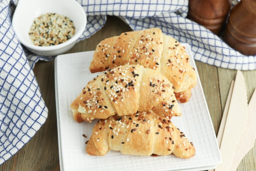 Plate of flaky everything bagel crescent rolls stacked on a white serving plate and topped with sesame seeds, poppy seeds, garlic, and onion seasoning.