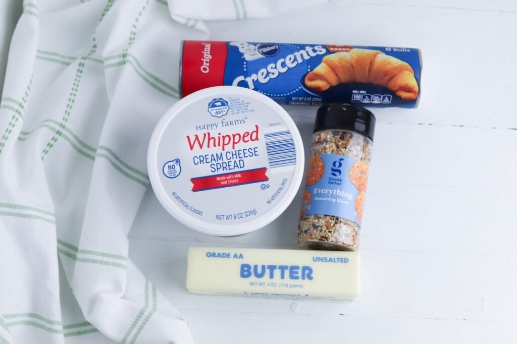 Ingredients for everything bagel crescent rolls including crescent roll dough, whipped cream cheese, butter, and everything bagel seasoning on a white surface.