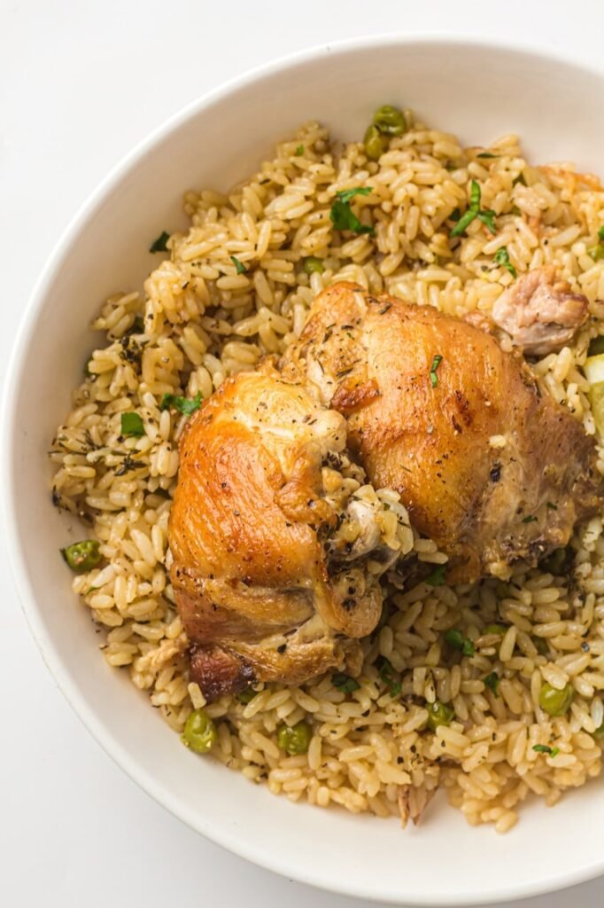 Bowl of Instant Pot chicken and rice topped with two golden-brown chicken thighs, fluffy seasoned rice, green peas, and chopped herbs, showing the finished one-pot chicken and rice dinner.
