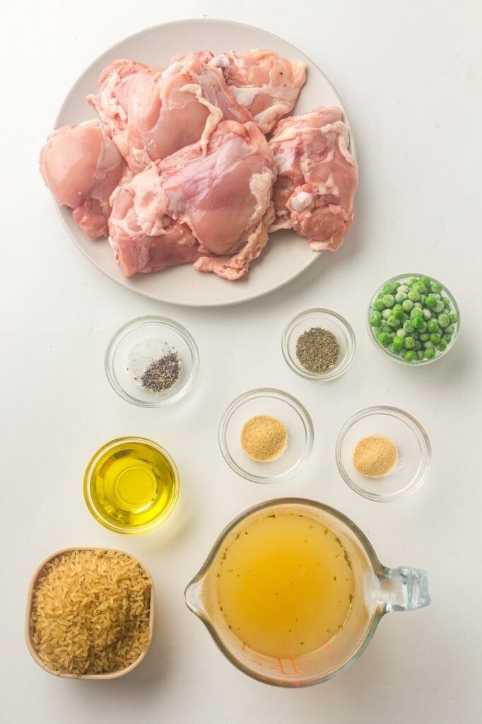 Ingredients for Instant Pot chicken and rice arranged on a white surface, including raw bone-in chicken thighs, rice, chicken broth, olive oil, seasonings, and frozen peas.