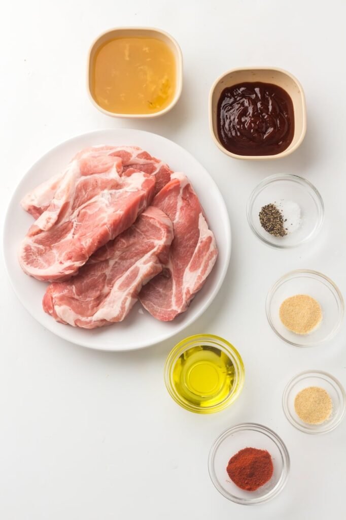 Raw pork shoulder steaks on a white plate surrounded by small bowls of olive oil, smoked paprika, garlic powder, onion powder, salt, pepper, chicken broth, and BBQ sauce for Instant Pot pulled pork.
