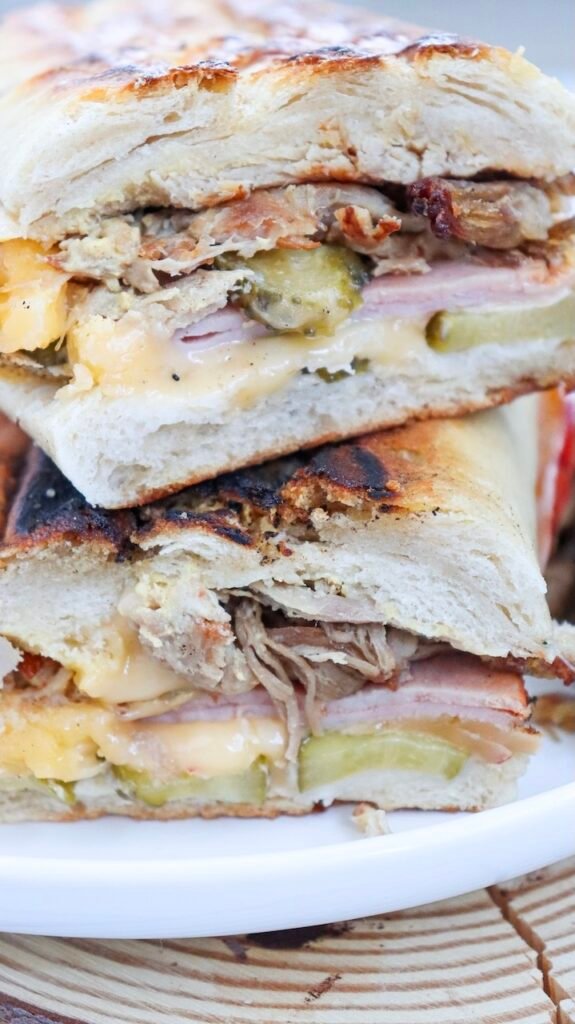 Close-up cross-section of a toasted Cuban sandwich stacked on a plate, showing layers of juicy roast pork, ham, melted cheese, and sliced pickles inside crispy golden bread.
