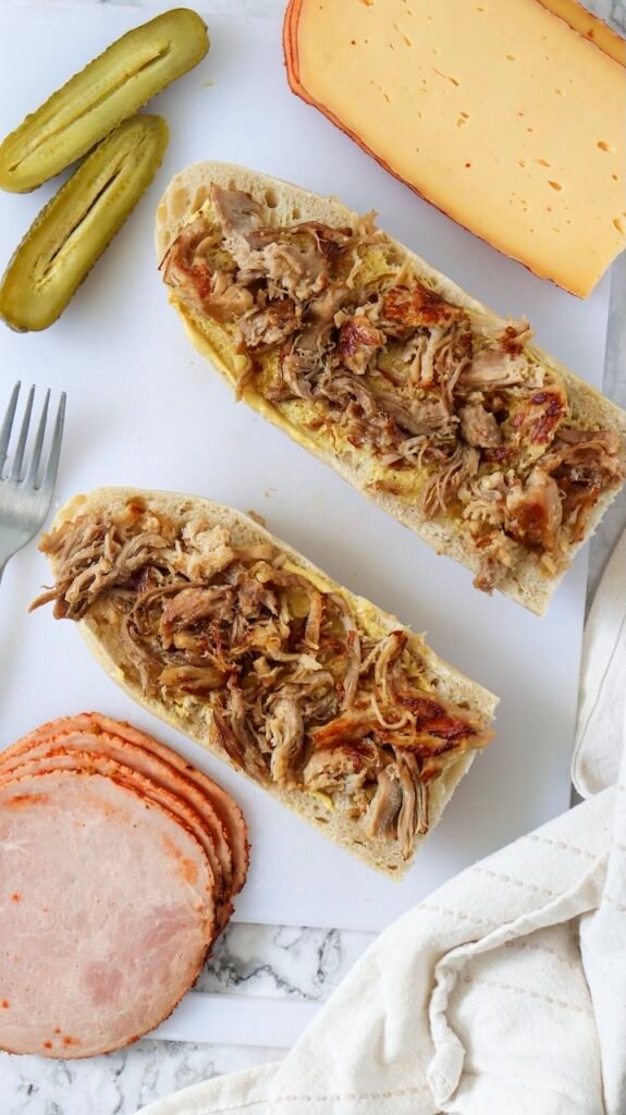 Overhead view of sliced Cuban bread spread with mustard and topped with shredded roast pork, with ham, Swiss cheese, and pickles nearby on a cutting board during Cuban sandwich assembly.