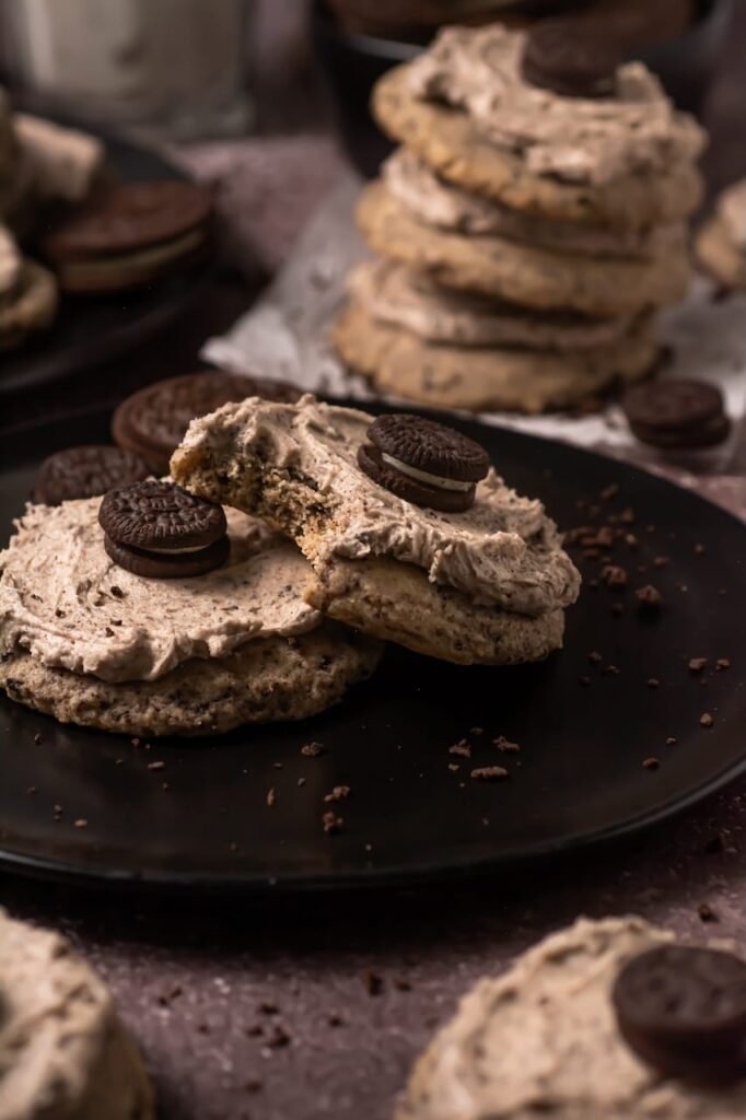 Close view of thick cookies cream milkshake cookies topped with Oreo frosting mini Oreo cookies on black plate, one cookie broken showing soft chewy interior.