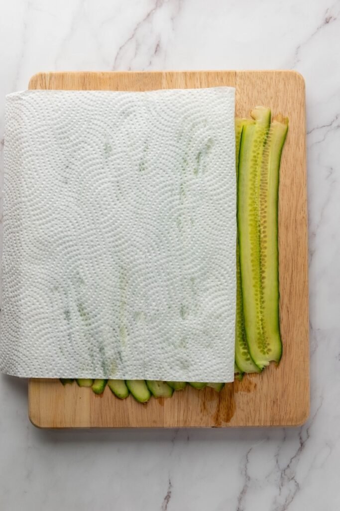 Paper towels placed over thin cucumber ribbons on a cutting board to remove excess moisture before assembling smoked salmon cucumber rolls.