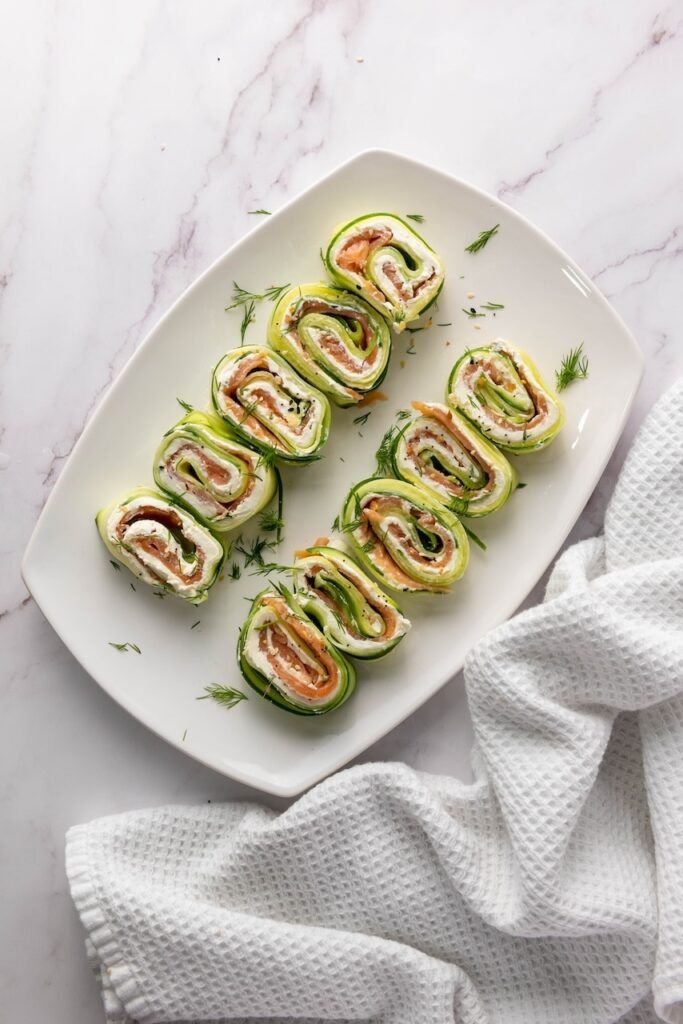 Overhead view of smoked salmon cucumber rolls arranged on a white plate, showing spiral layers of cucumber ribbons, cream cheese, smoked salmon, fresh dill, and everything bagel seasoning.