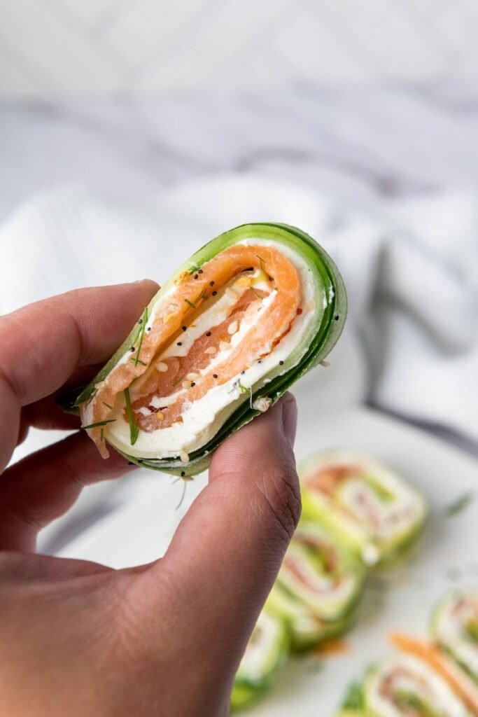 Hand holding a bite-sized smoked salmon cucumber roll showing layers of cucumber ribbon, cream cheese, smoked salmon, dill, and everything bagel seasoning.