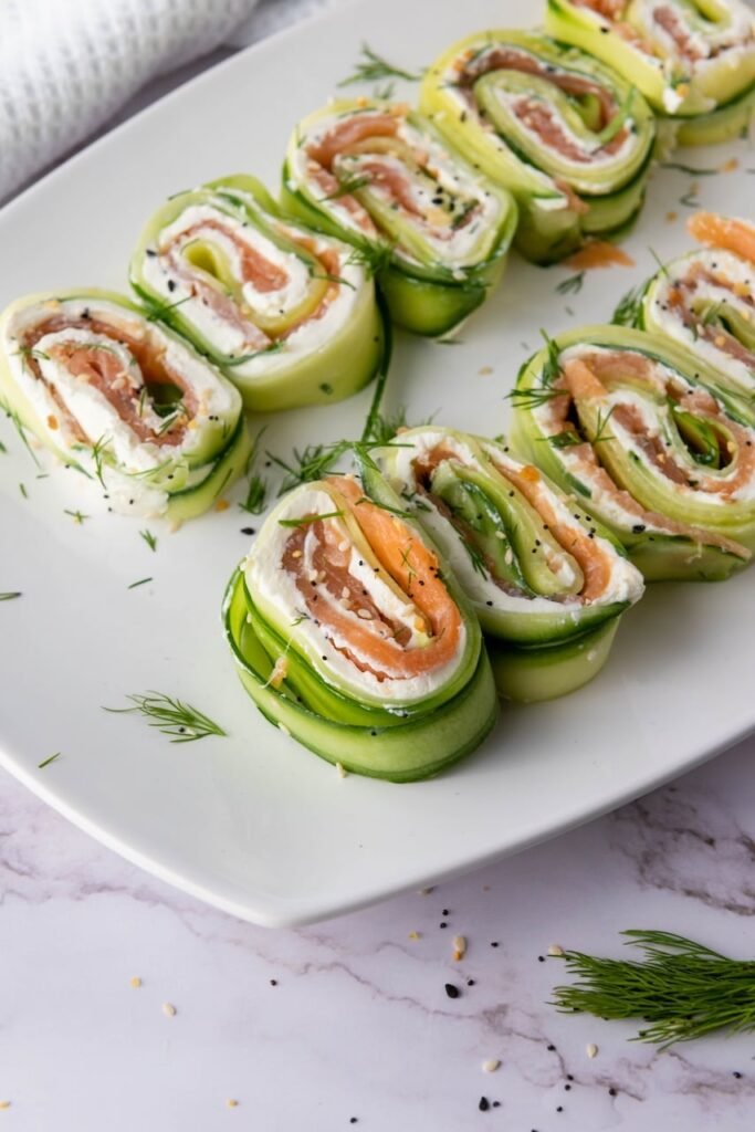 Close-up of smoked salmon cucumber rolls arranged on a white plate, showing layers of cucumber ribbons, cream cheese, smoked salmon, fresh dill, and everything bagel seasoning.