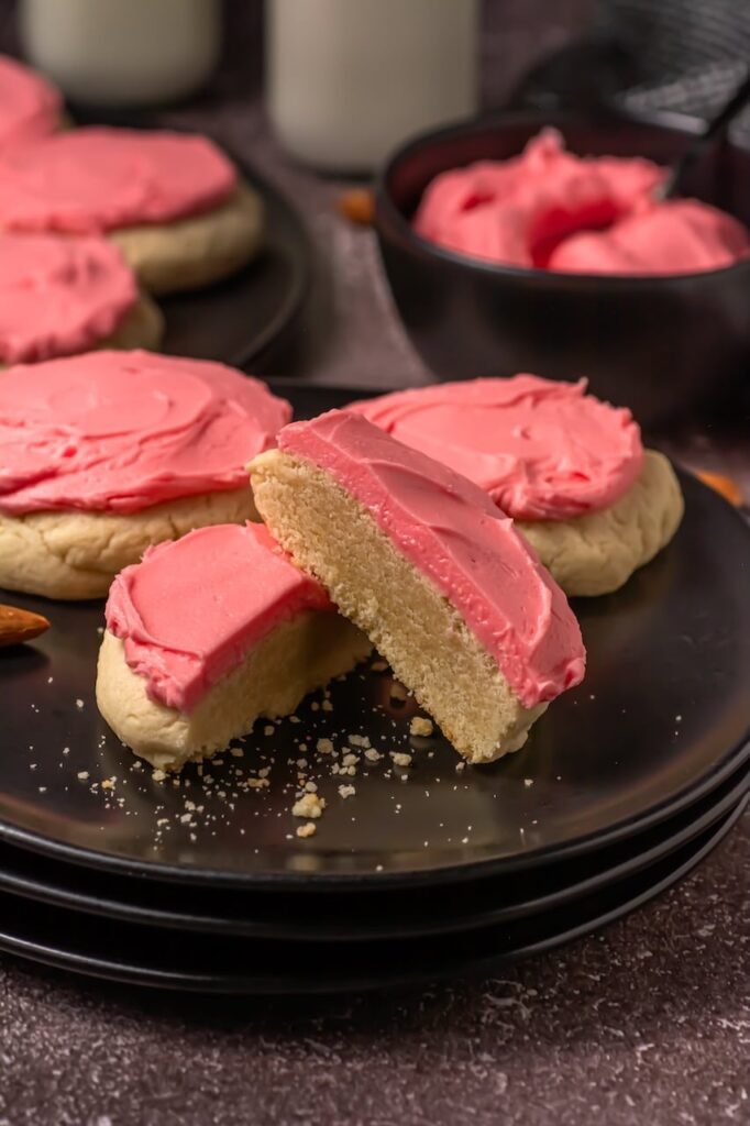 Close-up of soft sugar cookies with pink frosting arranged on black plates, styled with milk and almonds in the background for serving.