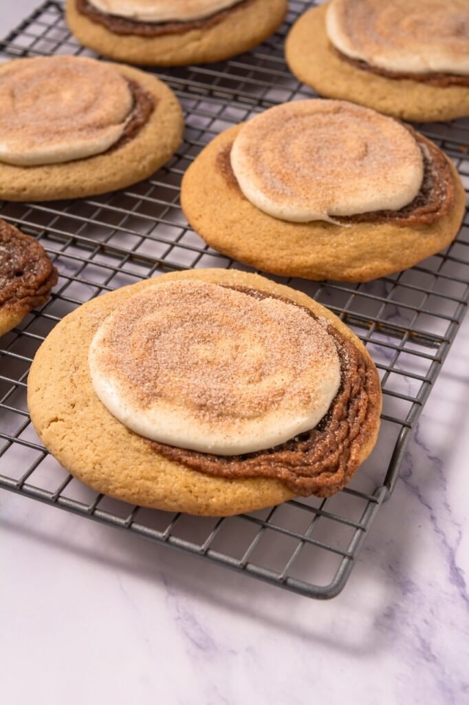 Close-up view of several baked Crumbl cinnamon swirl cookies resting on a wire cooling rack, each topped with cream cheese frosting and a light cinnamon sugar sprinkle, showing the thick cookie texture and cinnamon swirl layer.