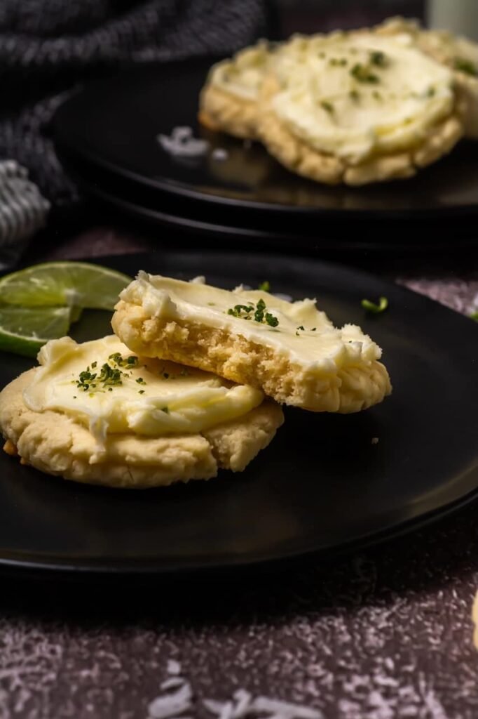Close-up of thick coconut lime cookies with creamy lime frosting, one cookie broken open to show the soft chewy center, served on a black plate with lime slices.