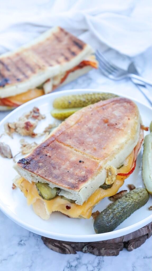 Toasted Cuban sandwich on a white plate with pickles, showing melted cheese and sliced pickles peeking out from crispy pressed bread.