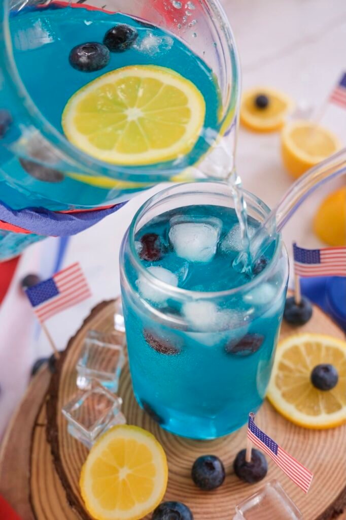 Blue lemonade mocktail being poured from a pitcher into a glass filled with ice, blueberries, and lemon slices for a 4th of July drink.