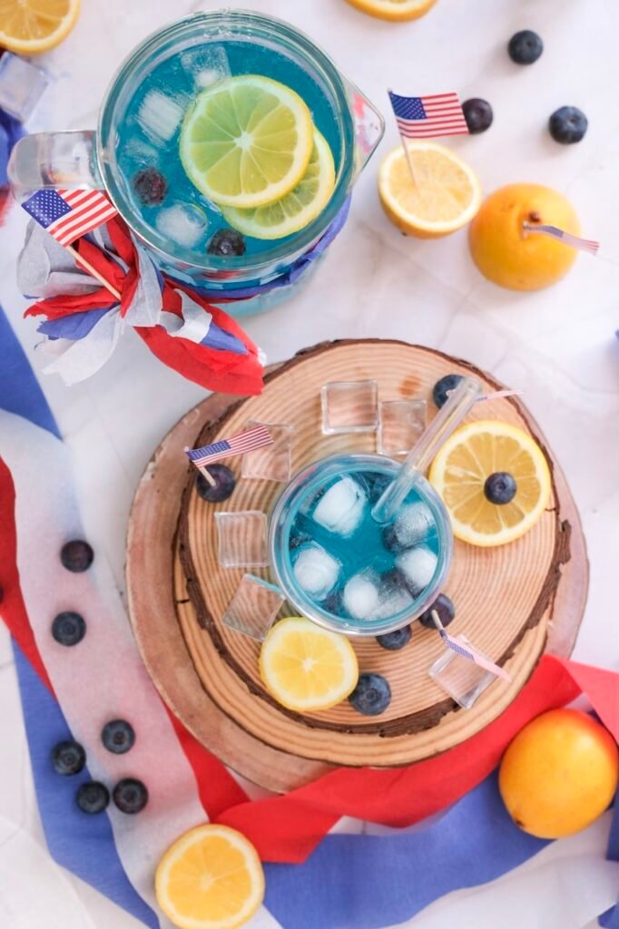 Overhead view of a blue 4th of July mocktail with ice, lemon slices, and blueberries, served in a glass and pitcher on a wooden board with patriotic decorations.