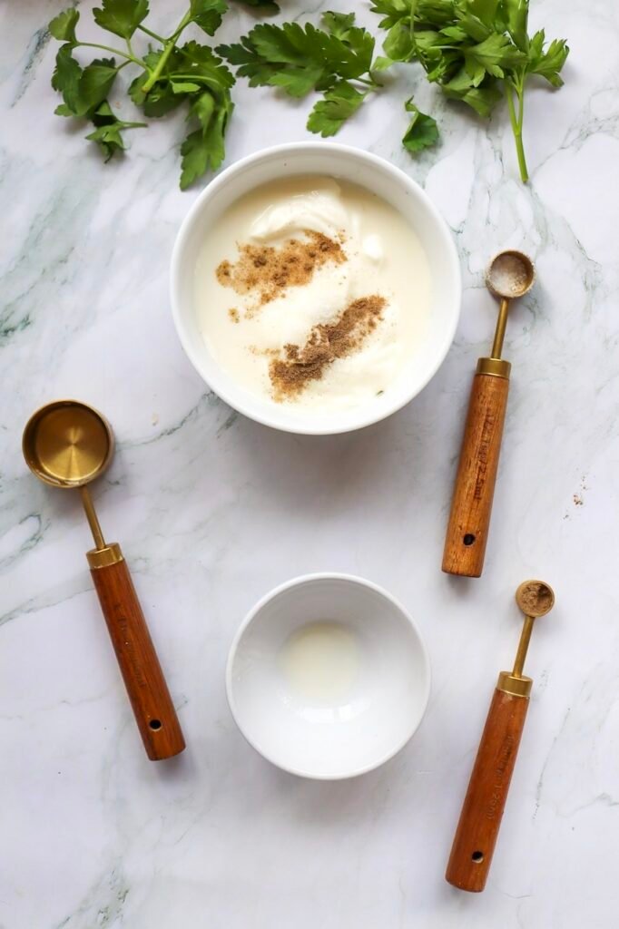 Overhead view of a bowl with mayonnaise, buttermilk, and spices being combined to make a creamy coleslaw dressing, with measuring spoons and parsley nearby.