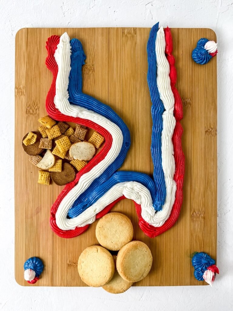 A wooden platter with red, white, and blue frosting decorated with cookies and snack mix, showing the step of assembling.