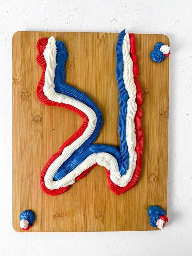 A wooden board with red, white, and blue buttercream frosting piped in decorative lines, showing the early design of a 4th of July dessert board before adding toppings.