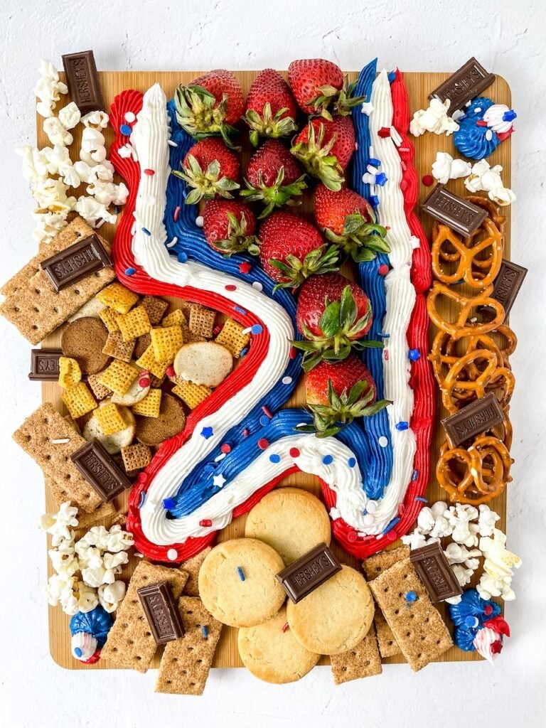 A finished snack with red, white, and blue frosting, fresh strawberries, cookies, pretzels, chocolate pieces, and popcorn arranged on a wooden.