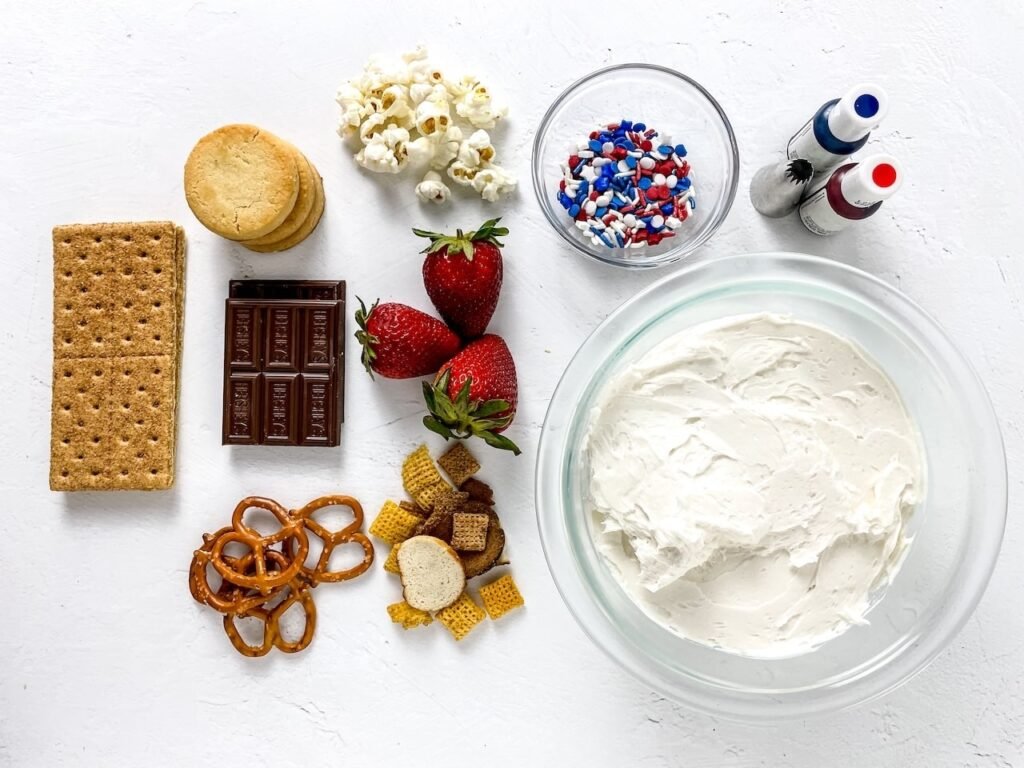 Overhead view of a bowl of white buttercream frosting surrounded by strawberries, pretzels, graham crackers, cookies, chocolate, sprinkles, and food coloring arranged on a white surface for a 4th of July buttercream board.