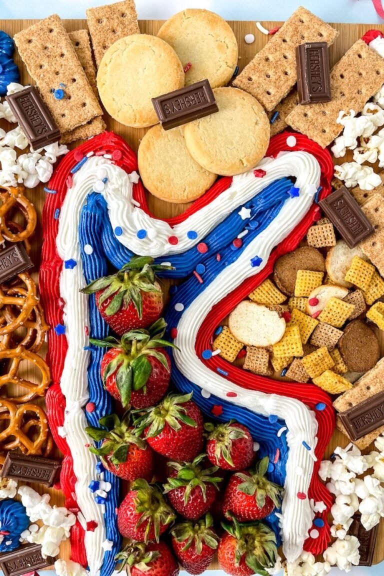 Close-up view of a 4th of July buttercream board with red, white, and blue frosting, strawberries, cookies, graham crackers, chocolate pieces, pretzels, and popcorn arranged on a wooden board.
