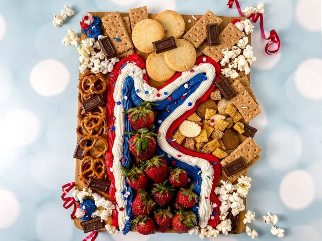 Top-down view of a patriotic dessert board with red, white, and blue buttercream frosting, strawberries, cookies, graham crackers, chocolate bars, pretzels, popcorn, and sprinkles on a wooden board with a soft blue background.