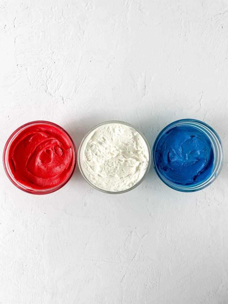 Three small glass bowls filled with red, white, and blue buttercream frosting arranged on a white background, showing the prepared frosting colors.